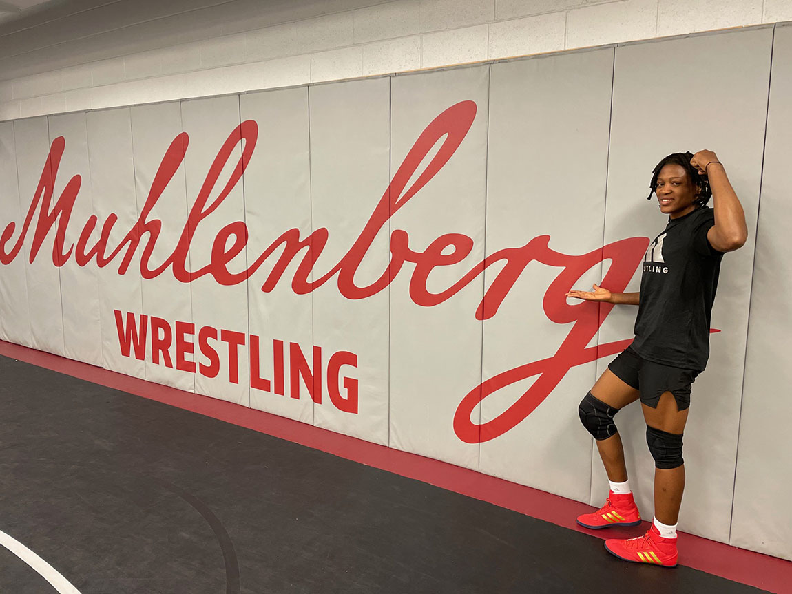 Odunayo Adekuoroye stands in front of a gym wall that says Muhlenberg WRESTLING