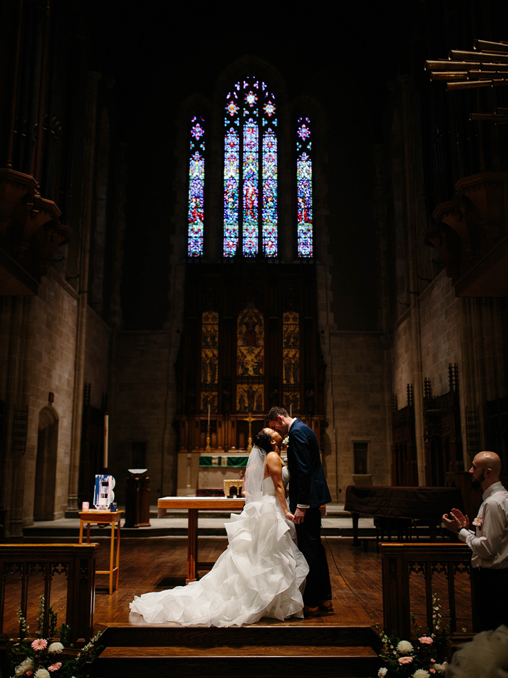 A groom, wearing a dark blue suit, leans in to kiss his bride, in a fluffy white wedding dress, inside a stone chapel with light streaming behind them from a stained glass window.