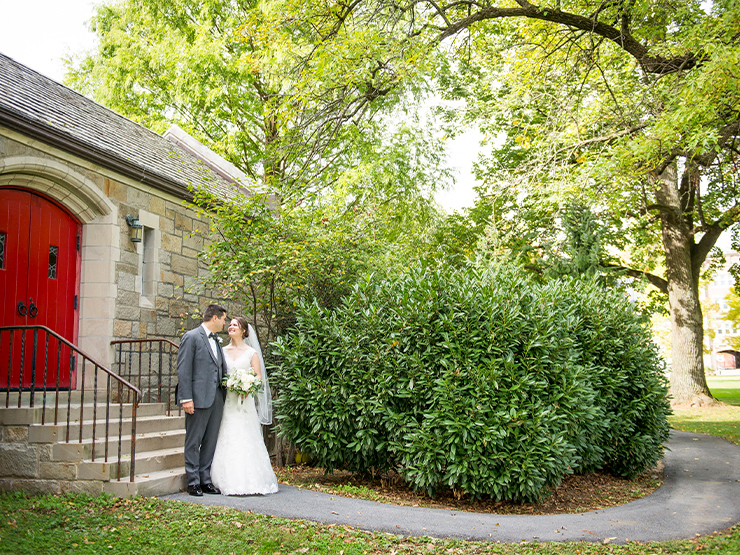 A bride and groom look into each others eyes at the side entrance of a stone chapel, surrounded by bright green foliage.