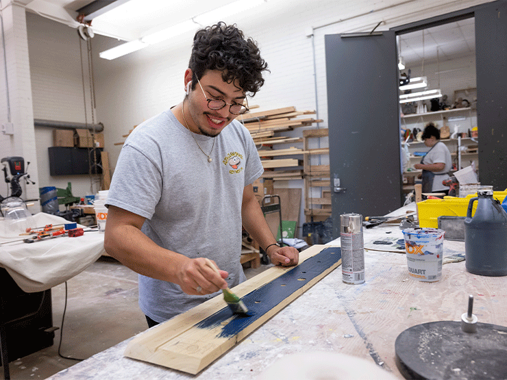 A student smiles while applying dark blue paint to a wooden board in an artist workshop.