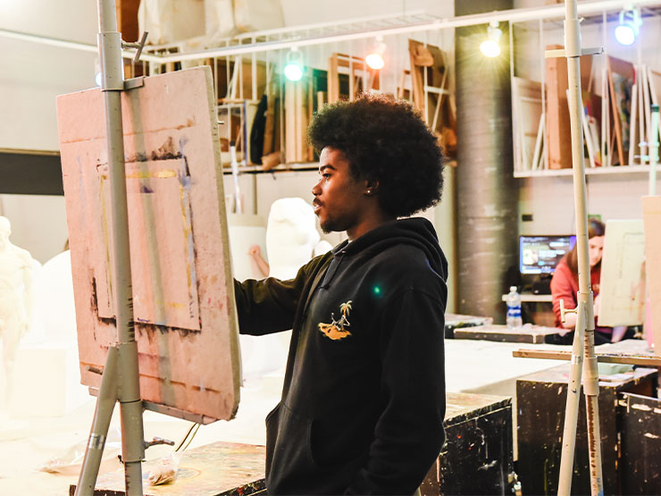 A student in a black hooded sweatshirt works with paints at an easel in an art studio.