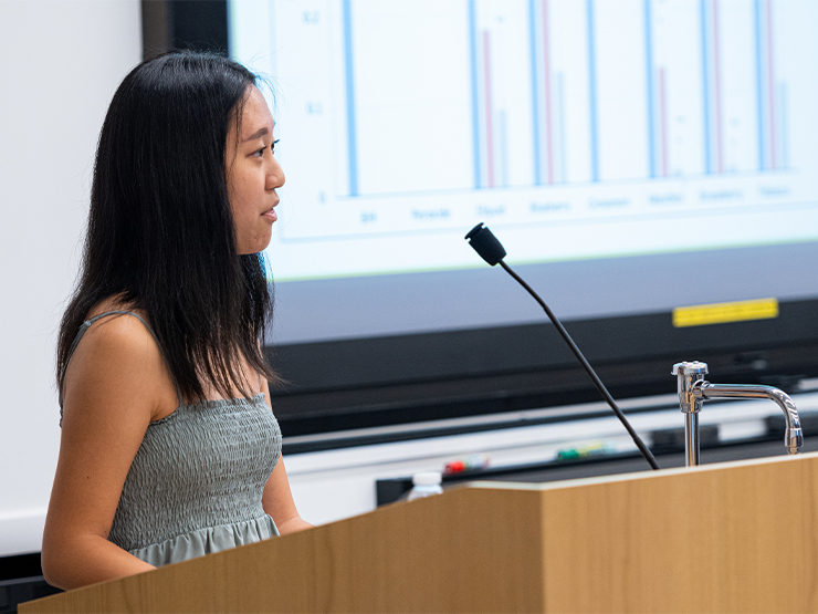 A young adult in a grey dress speaks at a podium in a classroom with data projected on a screen behind them.