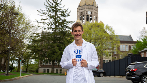 A white man with brown hair standing in front of the Haas Bell Tower in a white lab coat over a white Duke T-shirt