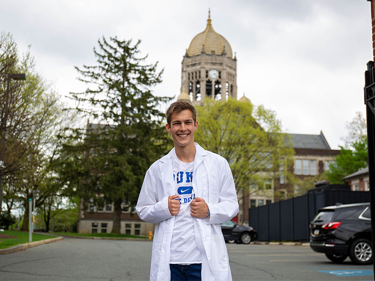 A white man with brown hair standing in front of the Haas Bell Tower in a white lab coat over a white Duke T-shirt
