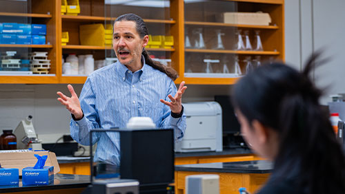 A professor with hair pulled back in a long ponytail speaks to students in a science lab.