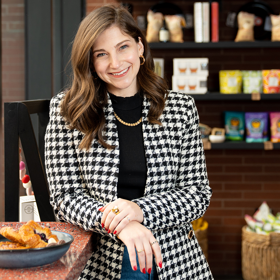 A smiling woman in a hound's-tooth jacket stands next to a table plated with pastries.