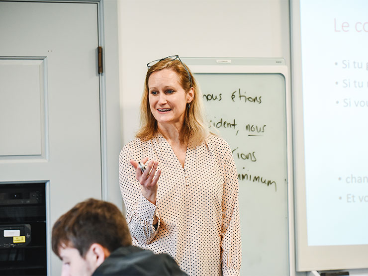An instructor dressed in a tan and black polka dot blouse speaks to a classroom while holding a dry erase marker.