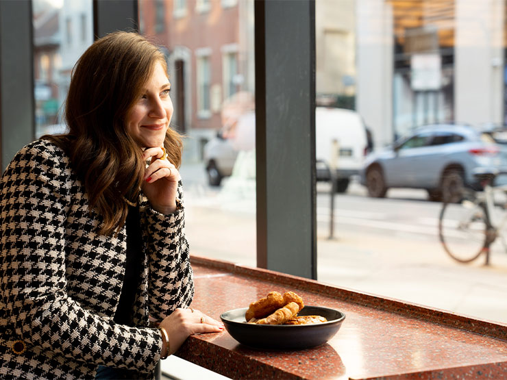 A woman in a hound's-tooth blazer looks out the window of a cafe with a bowl of pastries on the counter.