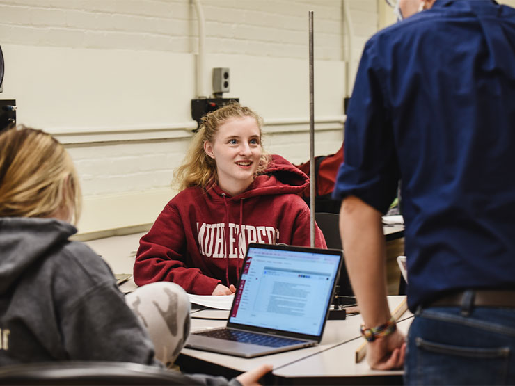 A student in a red Muhlenberg sweatshirt looks up at an instructor standing at a shared desk in a lab.