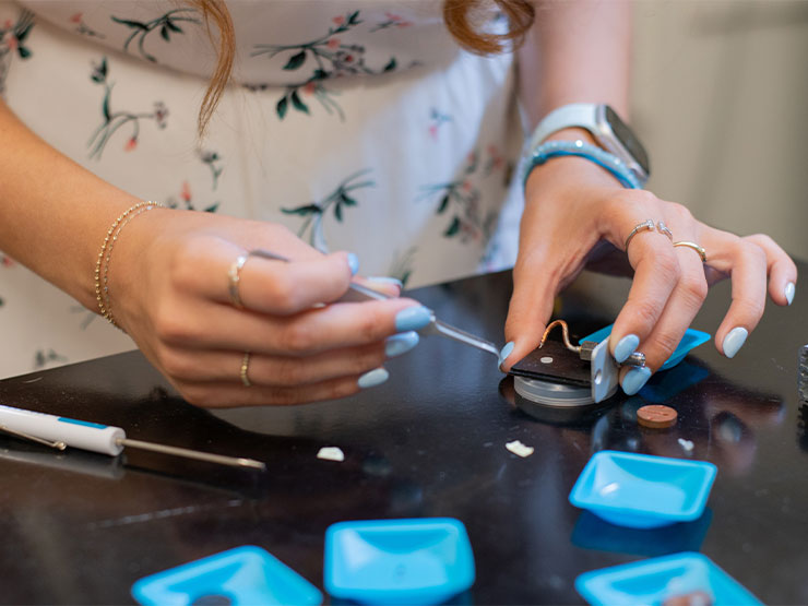 The hands of a student with light blue nail polish are seen working on equipment in a physics lab.