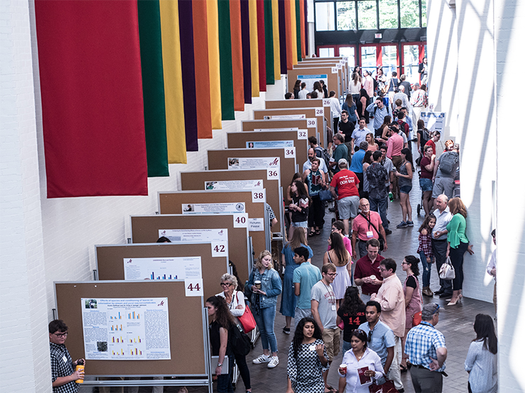 A large number of college students, faculty and staff peruse a research poster session in a large, open hallway with the Baker Center for the Arts.