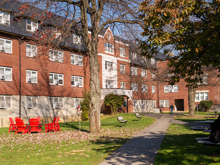 A brick residence hall stands against blue sky and autumn leaves.