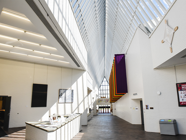 The lobby of the Baker Center for Arts, an open space with artwork and white cinderblock walls, is lit by sunshine from the peaked glass ceiling.