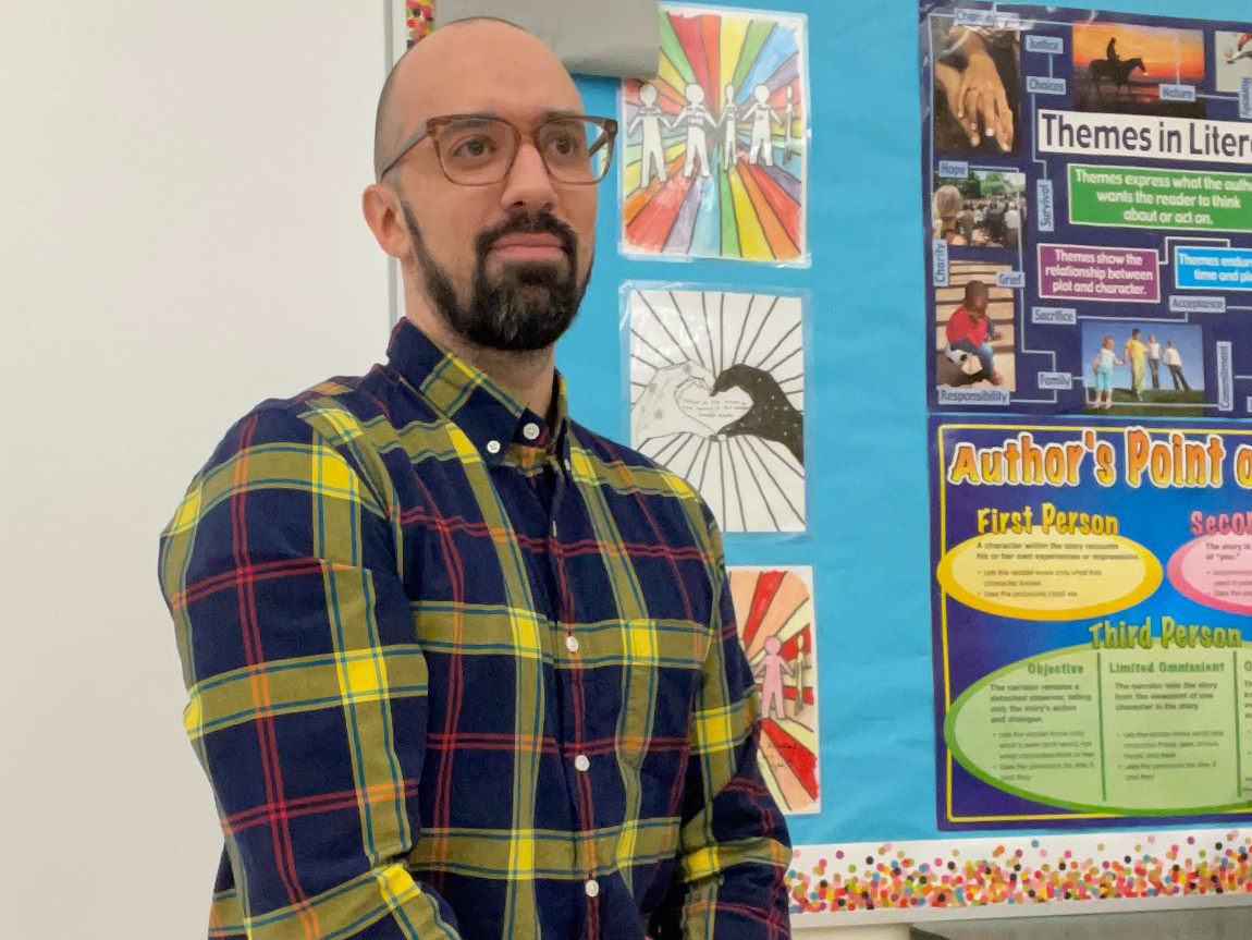 A bearded man sits in front of a classrooom bulletin board.