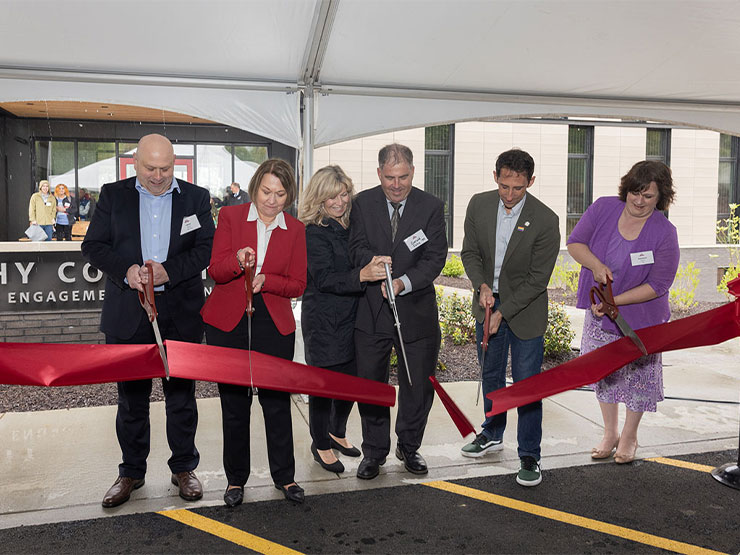 A group of six people uses giant scissors to cut a red ribbon in front of a new building