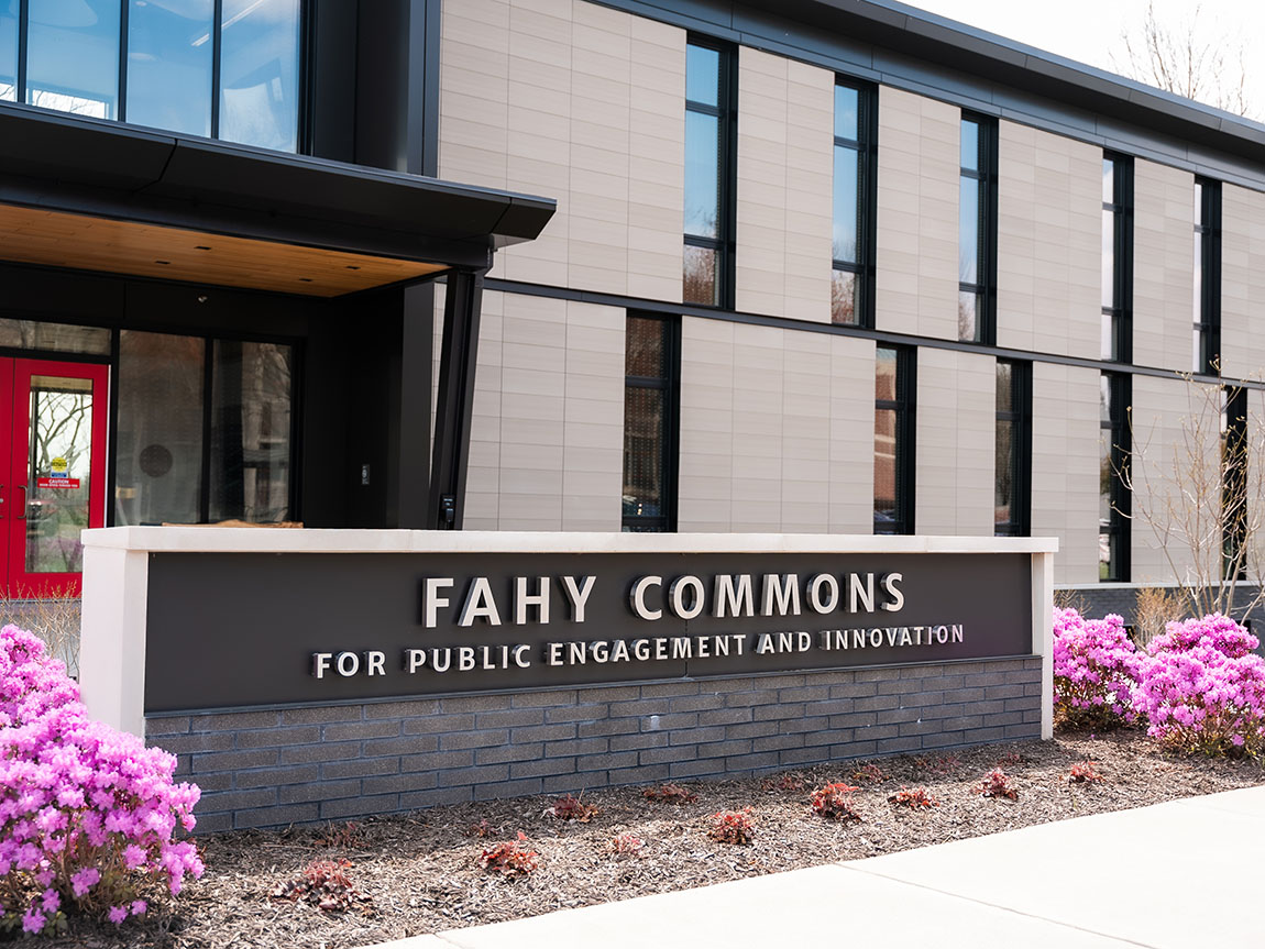 A sign in front of a building that says Fahy Commons and has purple flowering bushes on either side