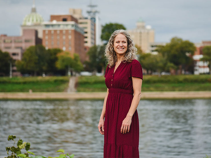 A woman with shoulder-length curly hair wearing a red dress smiles at the camera as she stands along the Harrisburg riverfront with the city skyline behind her.