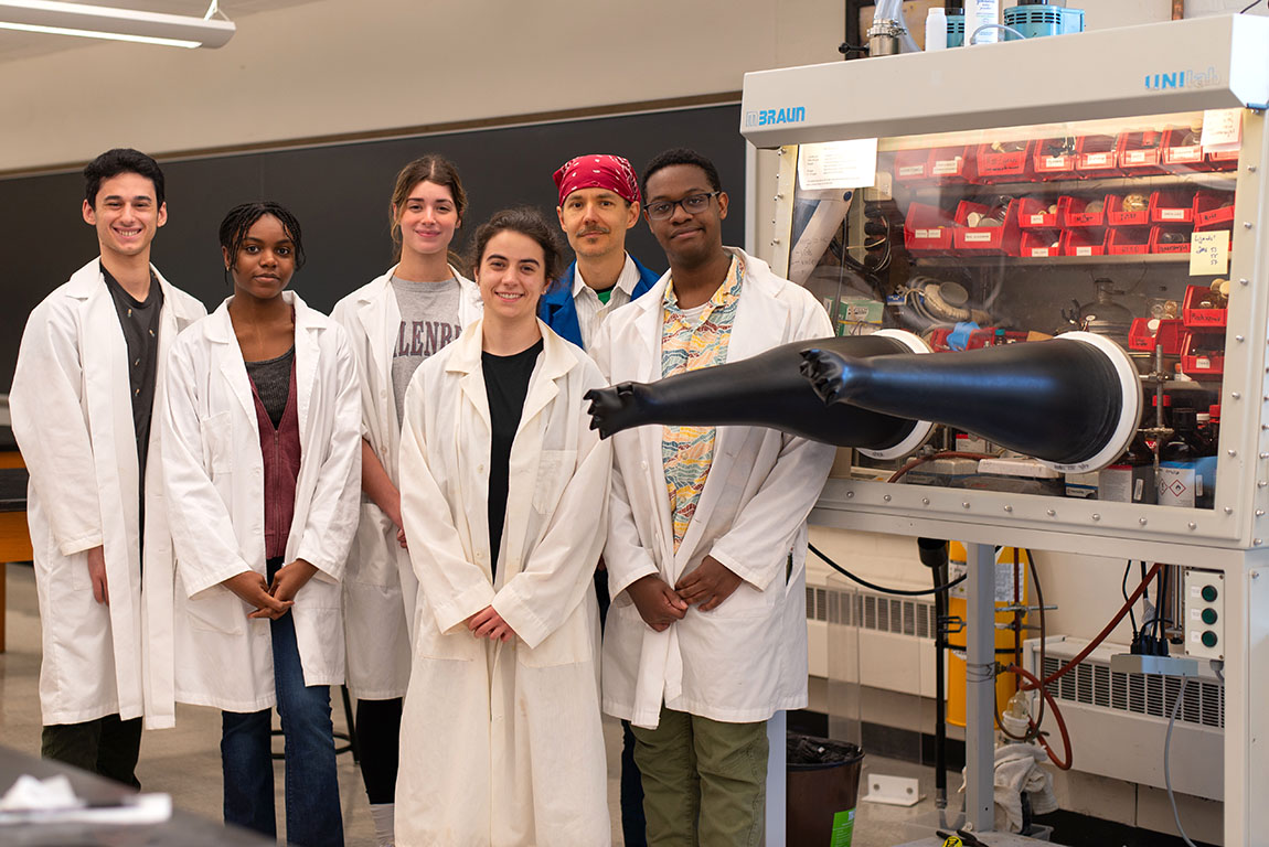 A group of college students and a college professor all wear white lab coats and pose next to a glove box in a chemistry lab