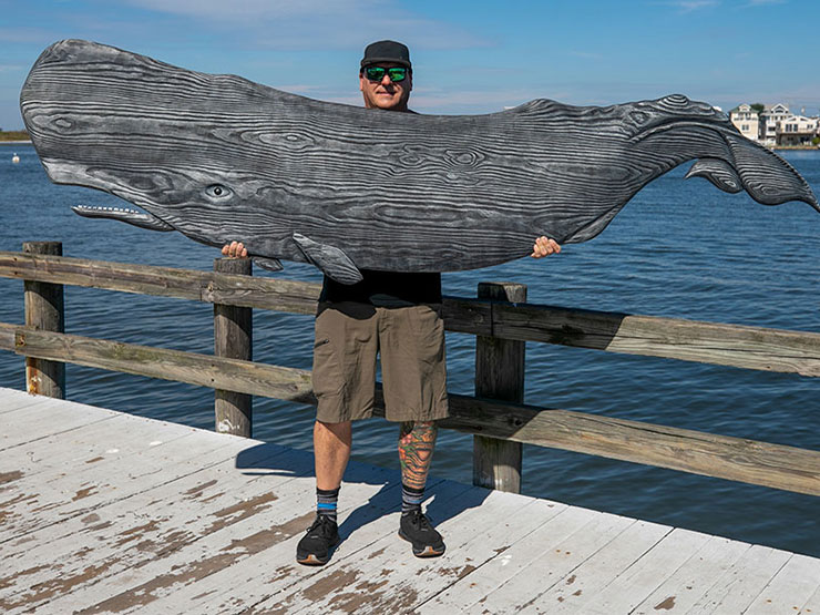 A man in a black baseball cap holds a giant wooden carved whale as he stands on a dock with water in the background