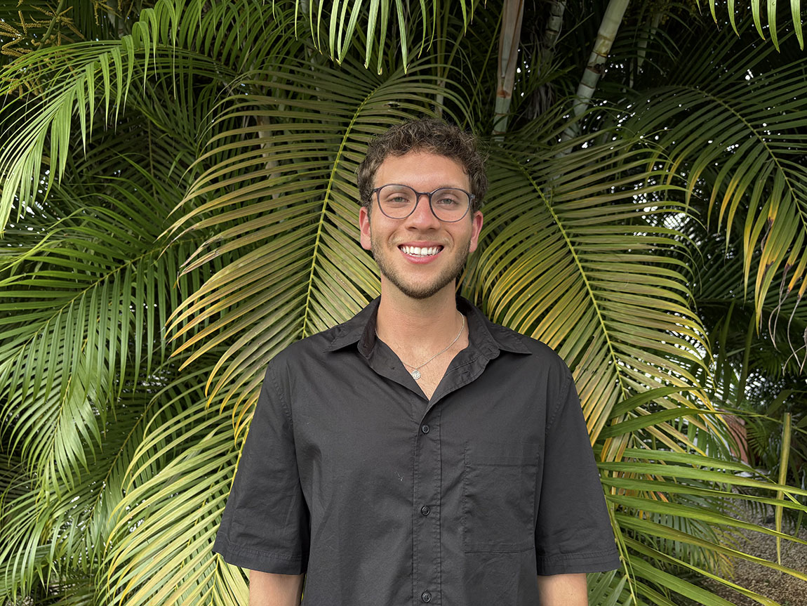 A college student in a black shirt and glasses stands in front of a bush