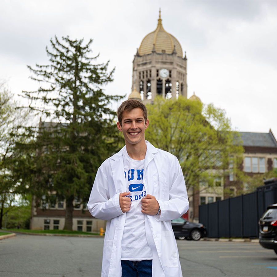 A white man with brown hair standing in front of the Haas Bell Tower in a white lab coat over a white Duke T-shirt.