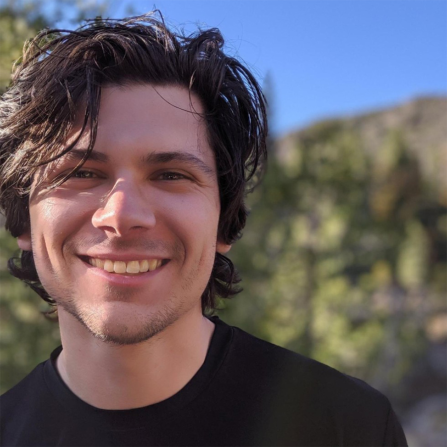 An adult in a black shirt smiles at the camera with an out-of-focus hilly landscape behind him.