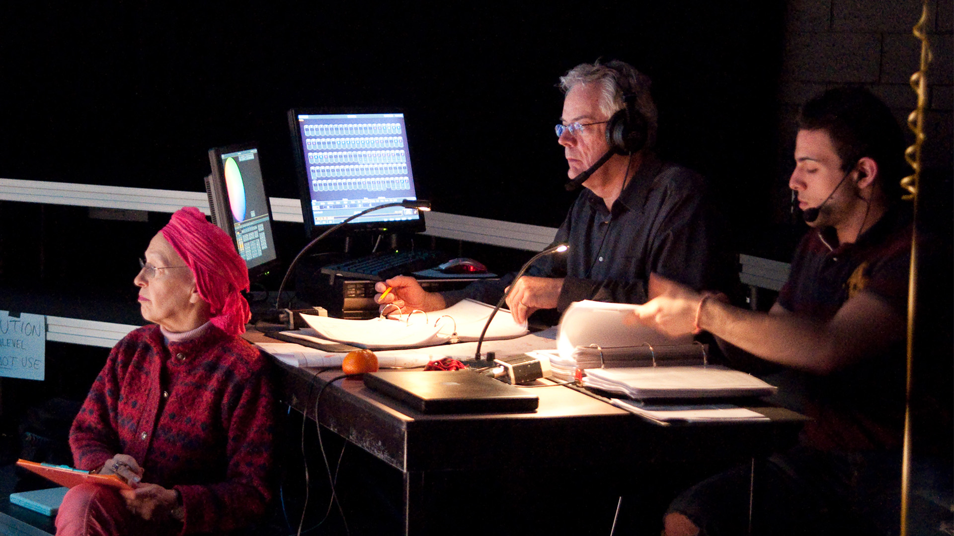 Members of the technical team for a production sit at a table in the Studio Theatre, watching a monitor and the stage.