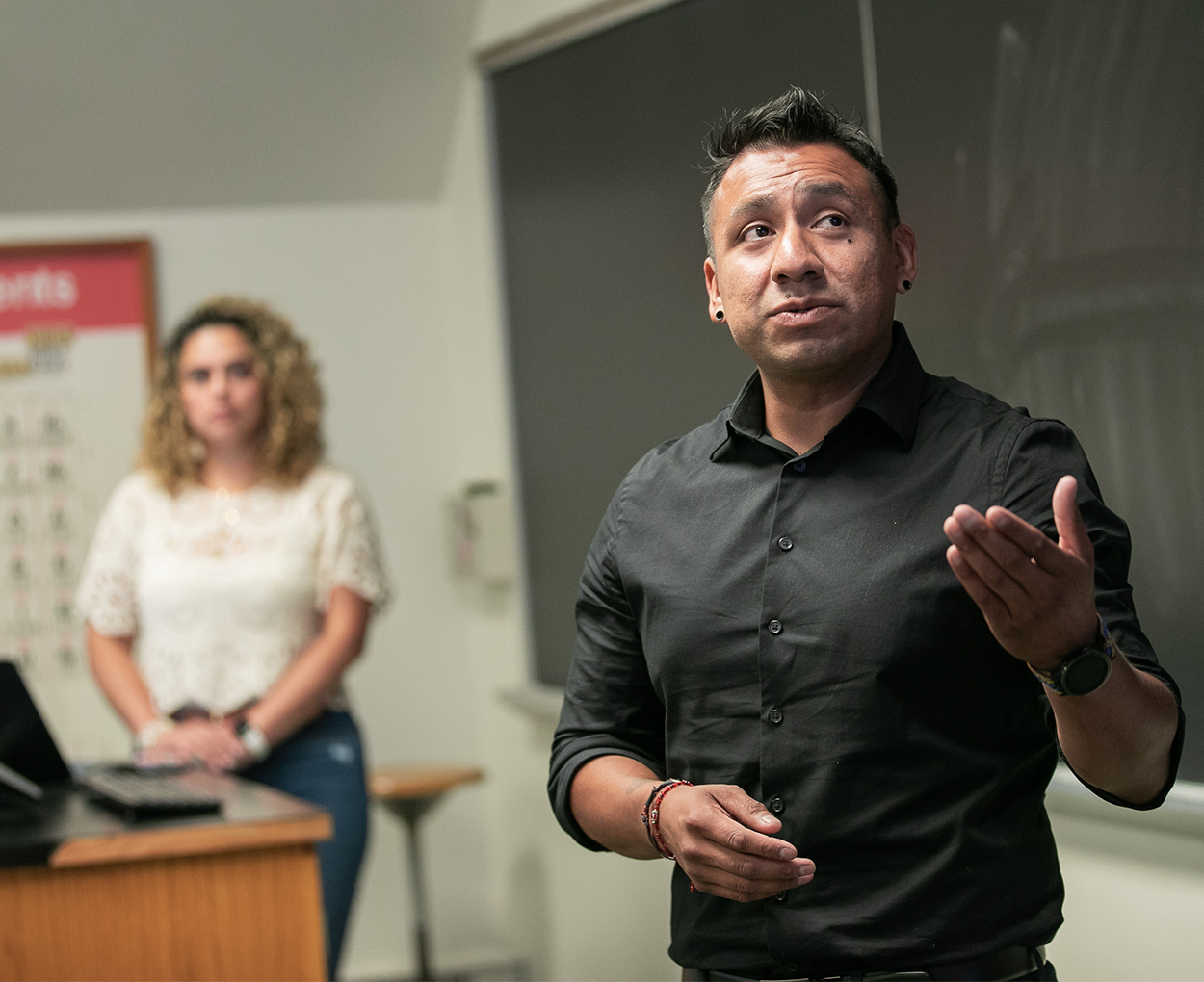 An adult in a black dress shirt speaks at a chalkboard while another adult standing at a desk with a computer looks on in the distance.