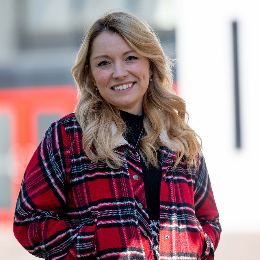 An adult with curly blonde hair, wearing a red flannel shirt, smiles at the camera.