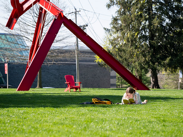 A student lies on the grass reading a book beneath the bright red, modern sculpture Victors Lament on the campus of Muhlenberg College.