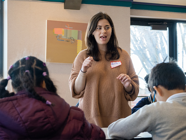 A female student wearing a name tag speaks to a group of elementary-age students in a classroom.