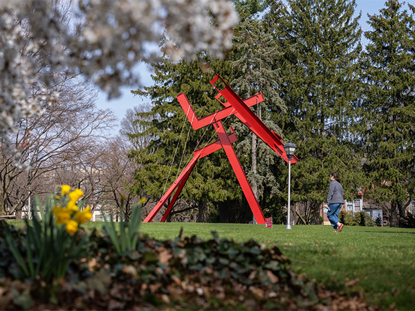 A large, bright red modern art sculpture stands on a college green, framed by newly budding spring trees.
