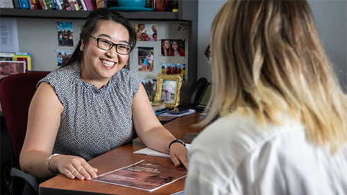 An adult at a desk in an office smiles while speaking with another person, who is slightly out of frame.