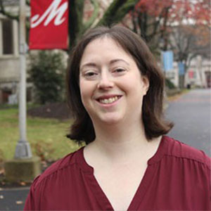 A smiling woman with short brown hair wears a maroon blouse while outdoors on the campus of Muhlenberg College.