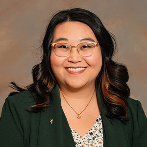 A photo of headshot of a smiling woman with curled dark hair, glasses and dark green blazer.