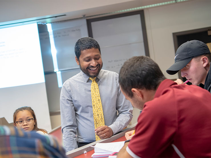 An adult in a yellow tie and dress shirt smiles while a student in a classroom works in front of him.