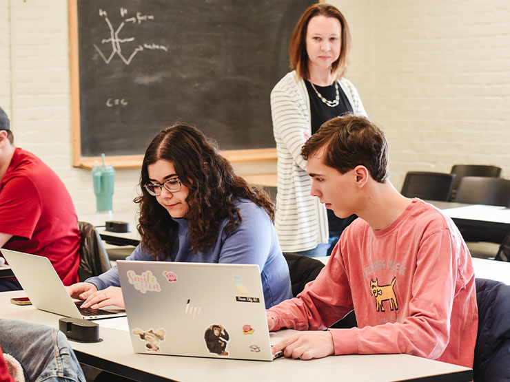 A college instructor paces a classroom, looking on as students work at laptops.