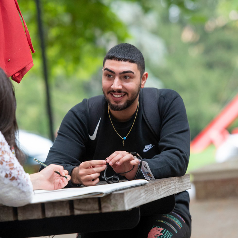 A student dressed in a black Nike shirt and pants sits at an outdoor picnic table and speaks with a person off-camera.