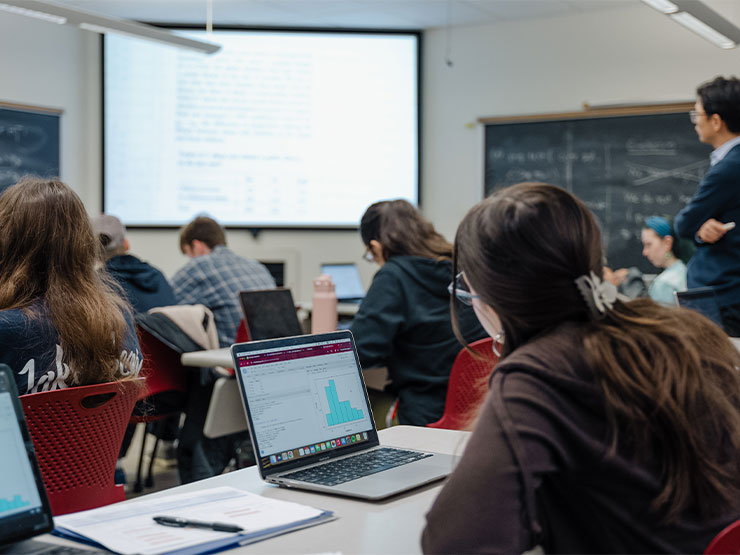 Students in a classroom with open laptops in front of them look toward a projector at the front of the room.