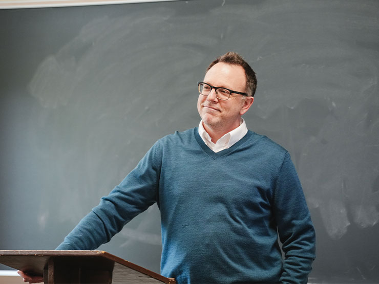 A college instructor in a blue sweater smiles while standing at a podium at the front of a classroom.