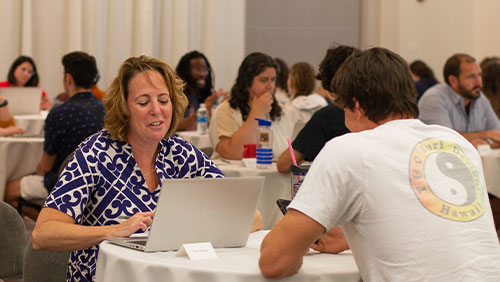 An adult looks at a laptop while sitting across the table from a young adult in a room crowded with such pairings.