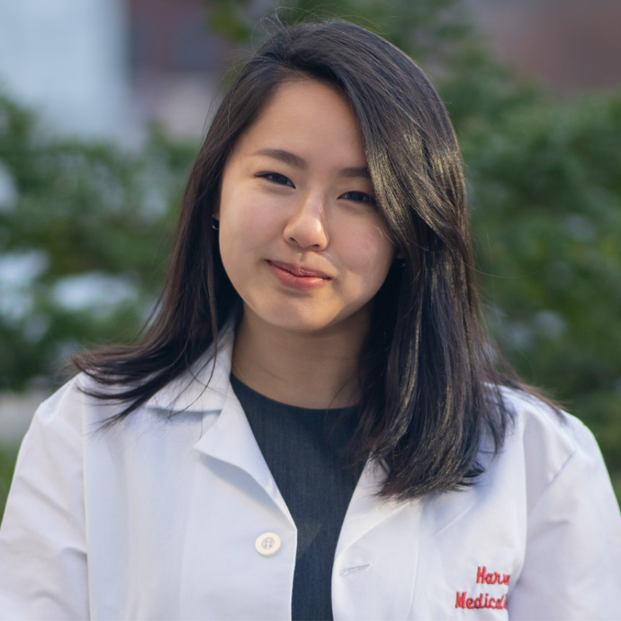 A student smiles at the camera outdoors while wearing a white lab coat embroidered with the words 