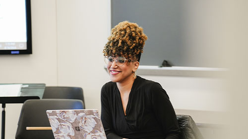 A woman wearing glasses and a black blouse smiles while seated in the front of a classroom.