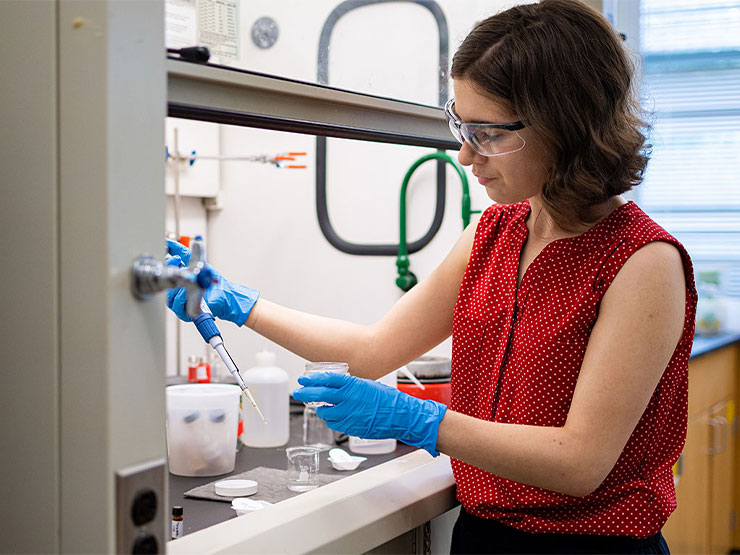 A college student, wearing gloves and goggles, works in a lab.