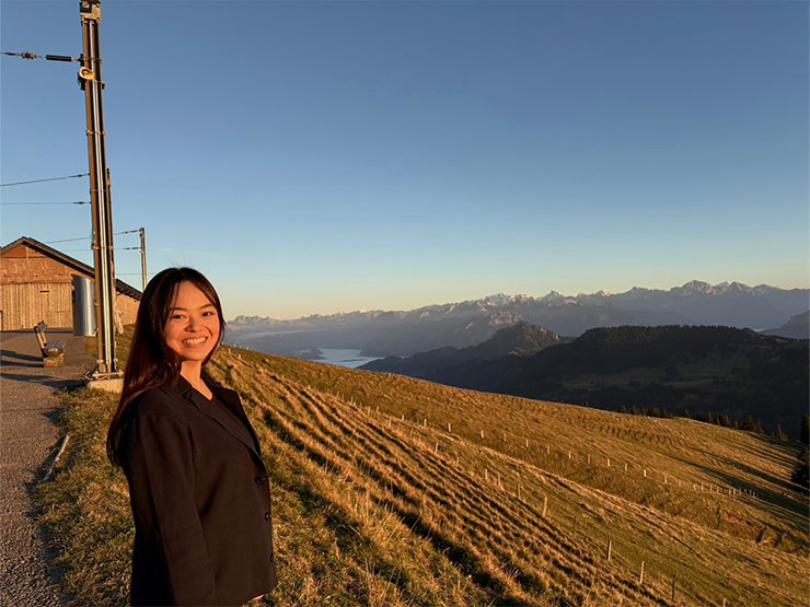 Fuka Aizawa stands smiling beside a field on a scenic vista in Switzerland.