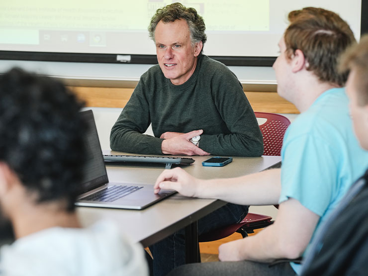 A college professor smiles and speaks to students seated around a table in a classroom.