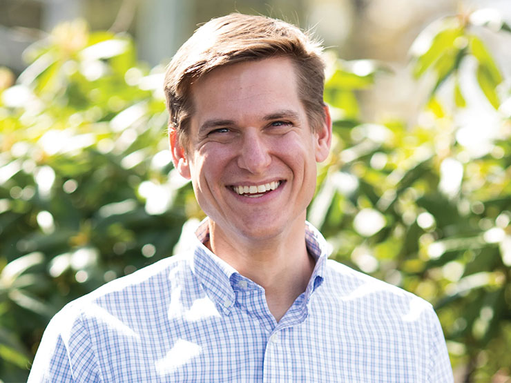 A smiling person in a blue and white checkered shirt smiles at the camera outdoors.