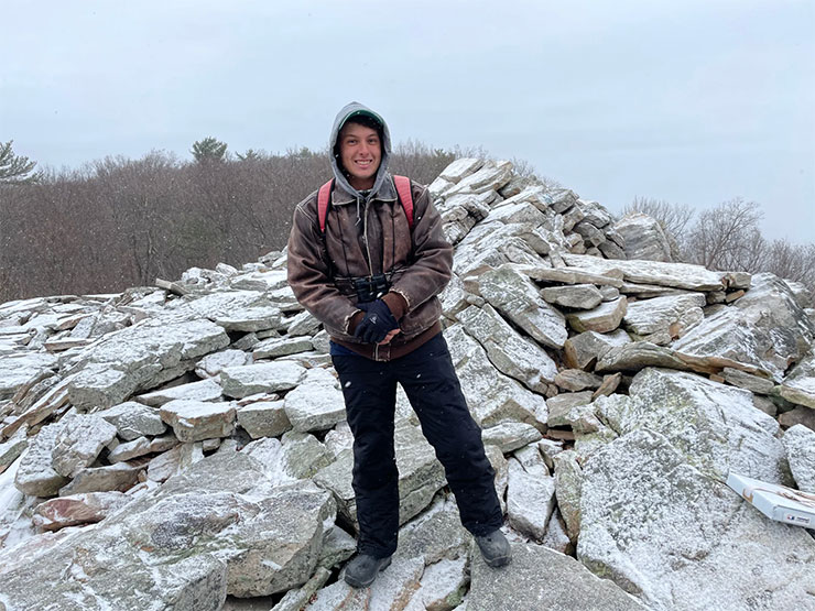 A young adult, dressed for cold weather, stands in a boulder field in a forest as snow falls.