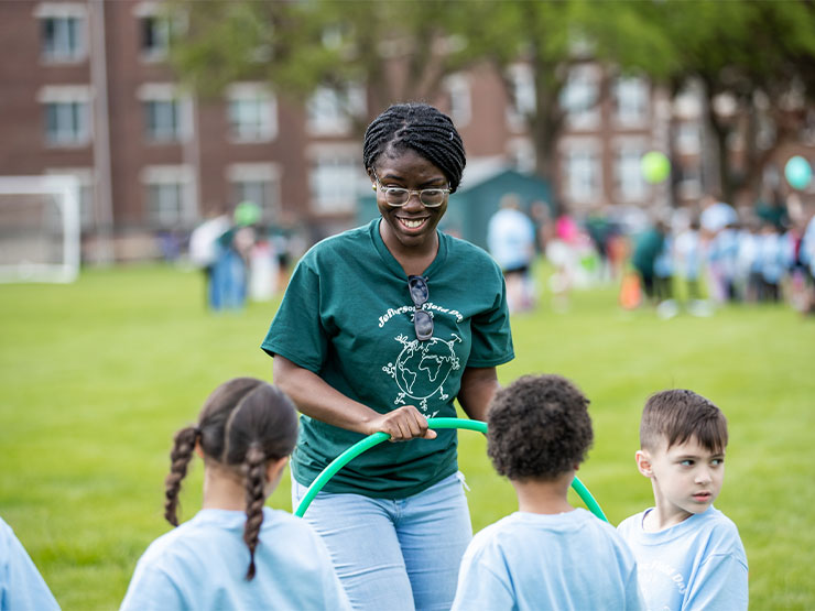A young adult in a green shirt speaks to elementary school grade children dressed in light blue shirts outside on a college campus.