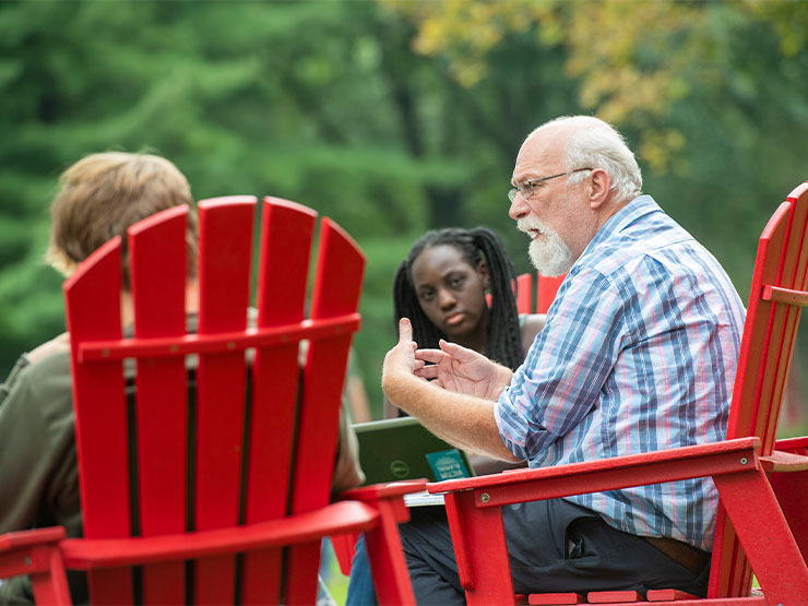 Students, seated in red Adirondack chairs, surround an instructor while holding class outdoors on the Muhlenberg College campus.
